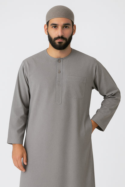 Man wearing an ash grey traditional thawb with round neckline, button placket, and chest pocket, standing against a light neutral background, showcasing modest Islamic menswear.